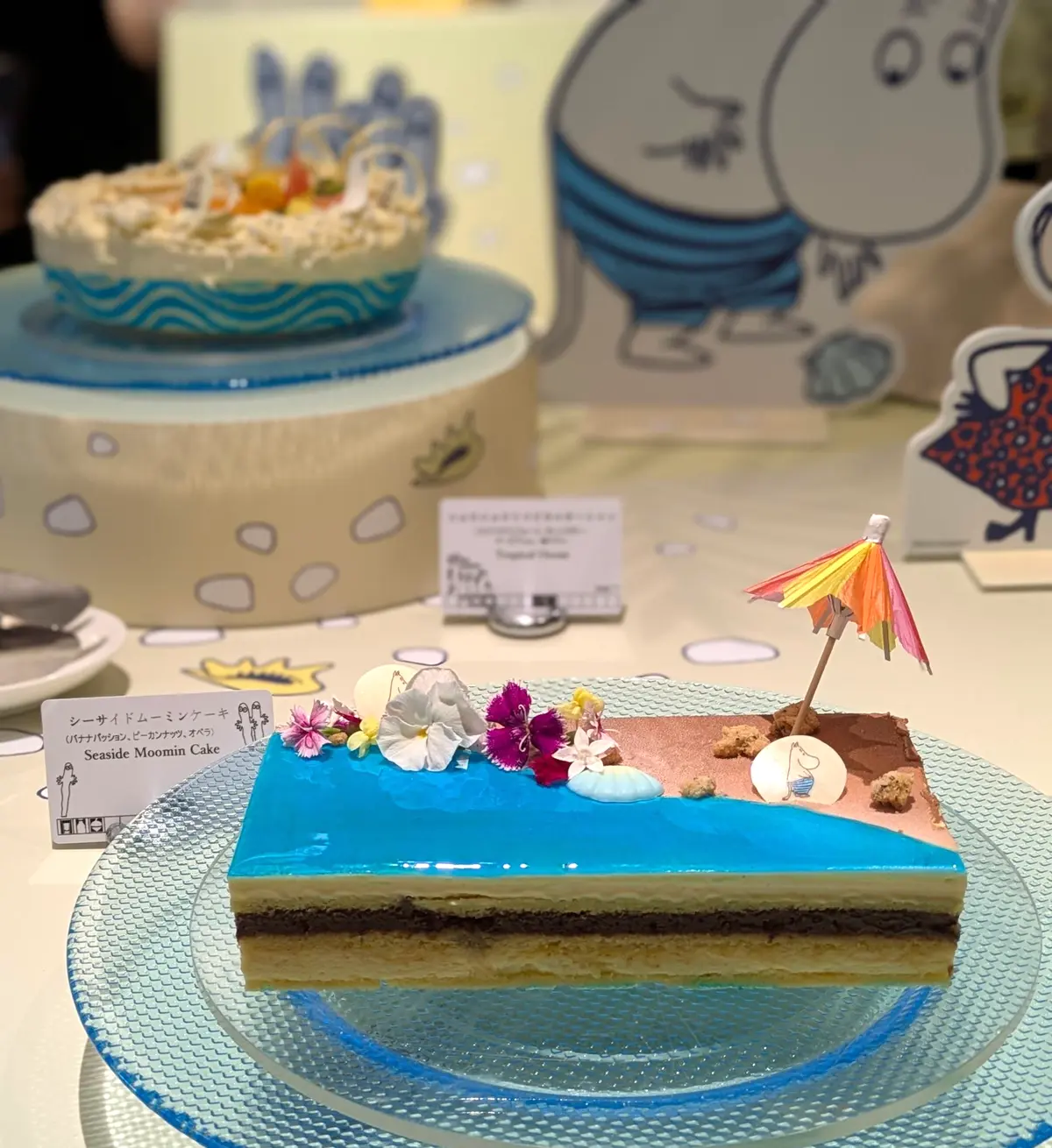 ビーチをテーマにしたケーキで、青い海を模したクリームの上に白い花、小さな傘、砂浜の装飾が置かれている