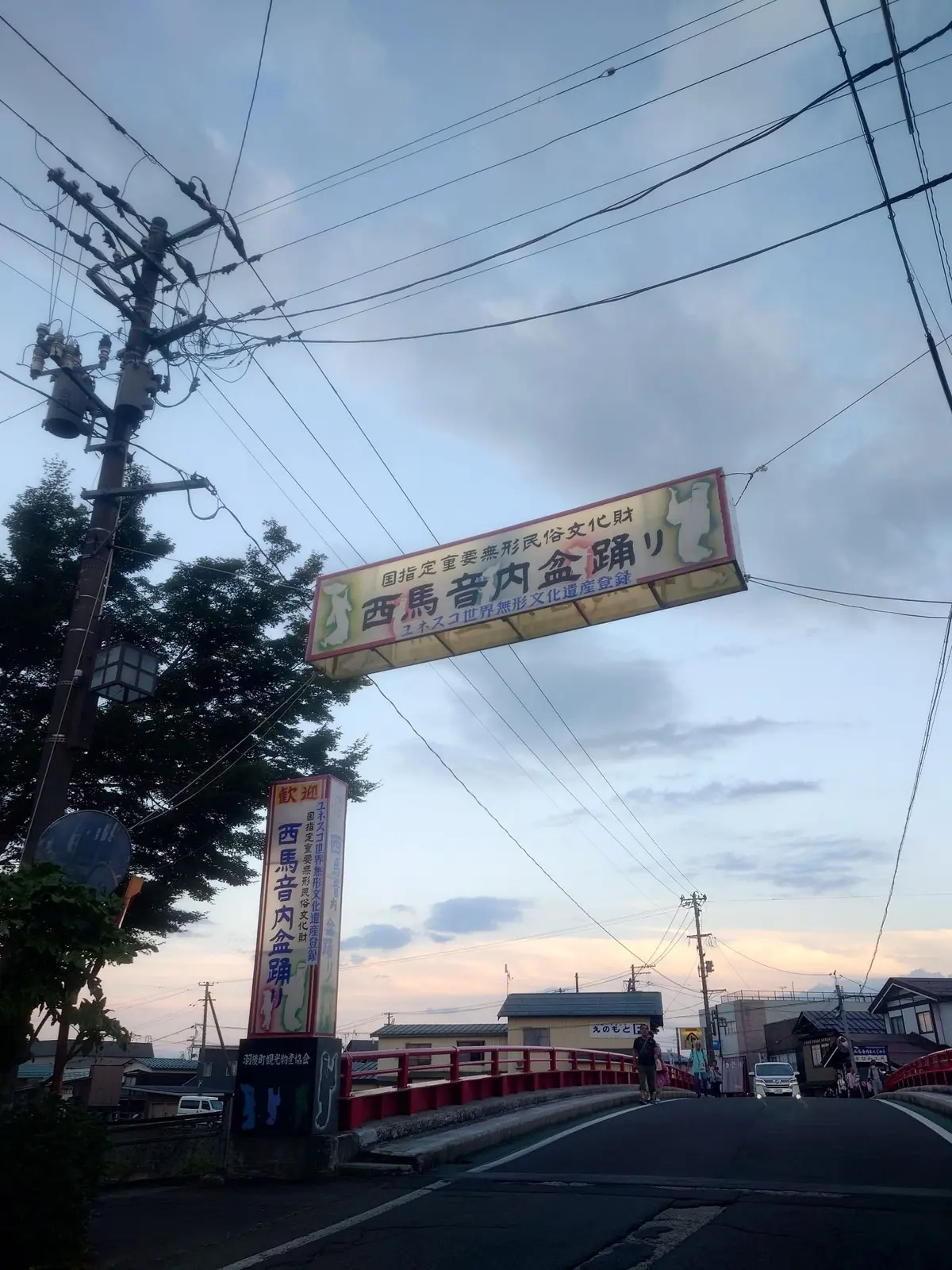 夕暮れ時の道路脇に設置された、黄色と茶色の看板。看板には日本語で「西馬音内金銅器」と書かれており、国の重要無形民俗文化財に指定されていることを示している。電柱や旗が看板の周囲に立っている。