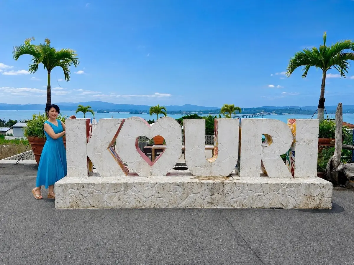 青いドレスの女性が「LOVE」の文字を背景に、海と山が広がる景色の中でポーズしています。