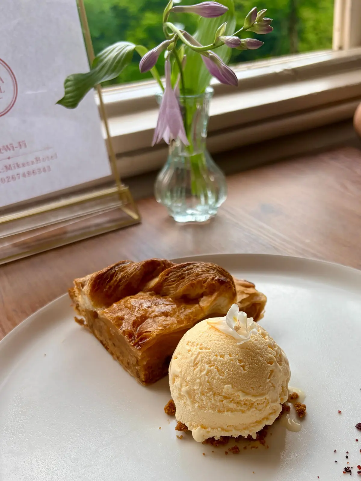 白い皿に、アップルパイとバニラアイスクリームが盛り付けられている。アップルパイは断面があり、中にはリンゴが詰まっている。アイスクリームは丸く、白いホイップるクリームが添えられている。背景には窓辺に置かれた花瓶と、メニューが置かれたイーゼルがある。