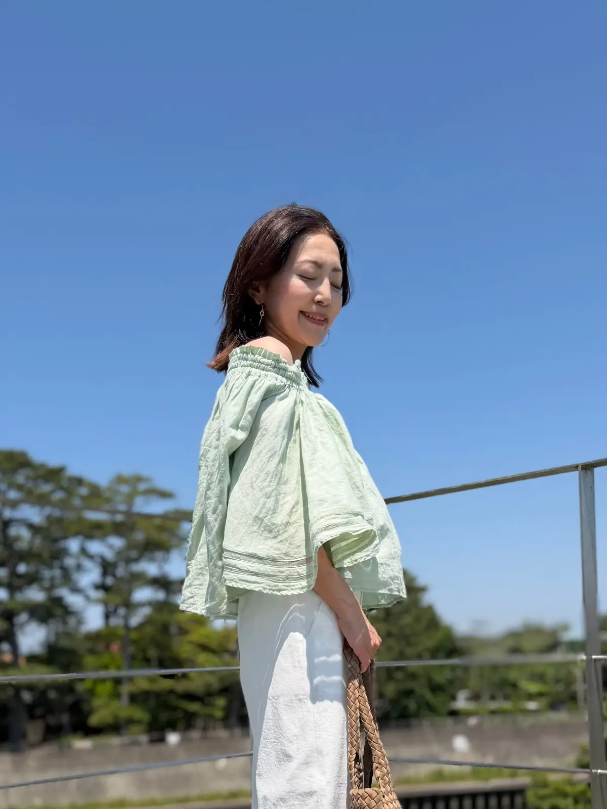 ミントグリーンのオフショルダートップと白のスカートを着た女性が、晴れた日に屋外で微笑んでいる。右手に編み込みのストラップバッグを持っている。背景には手すりがあり、木々がぼやけて見える。