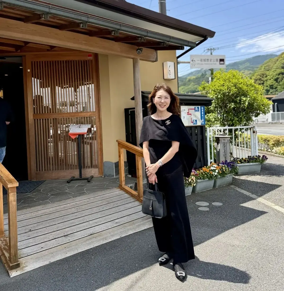 黒いワンピースに黒いショールを羽織った女性が、笑顔でポーズをとっている。背景には、木造の建物と駐車場、植栽が見える。