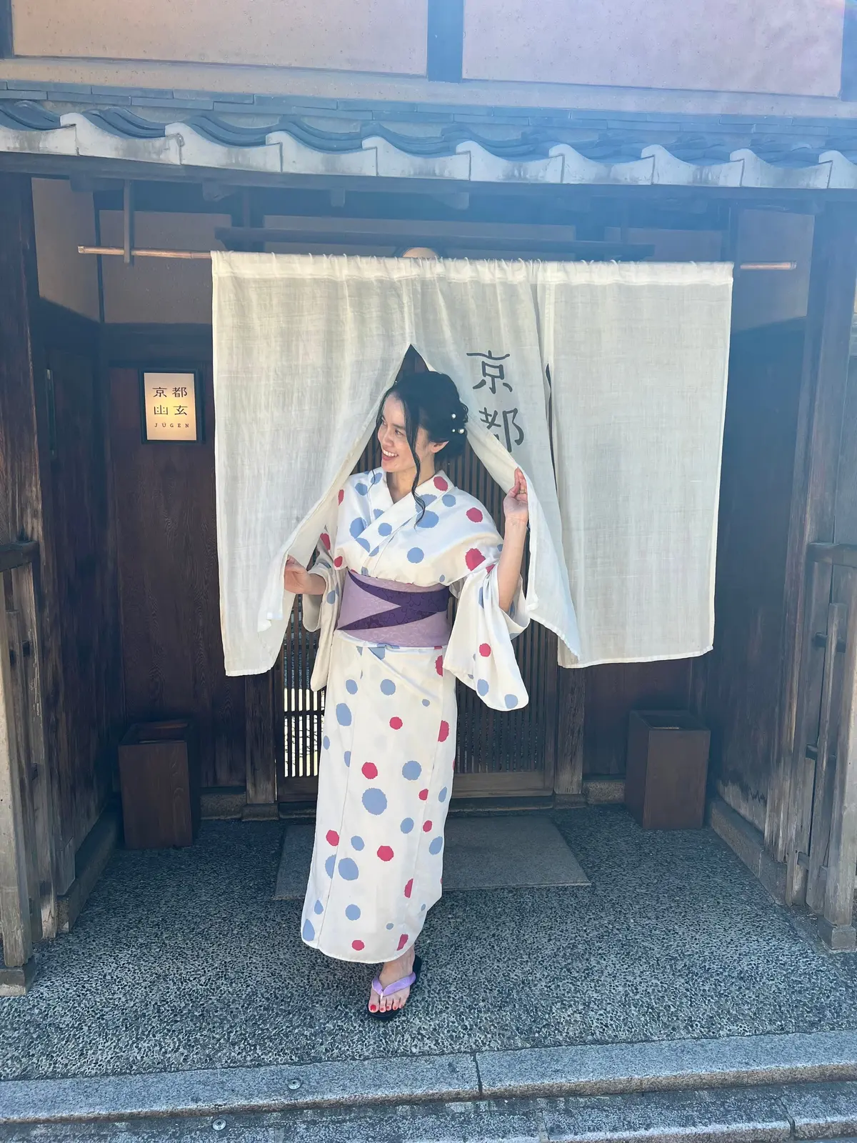白い壁と障子戸の前で、水色と白のドット柄の着物を着た女性がポーズを取っている。黒髪をまとめ、扇子を持っている。背景には日本語の文字が書かれた白い布がかかっている。