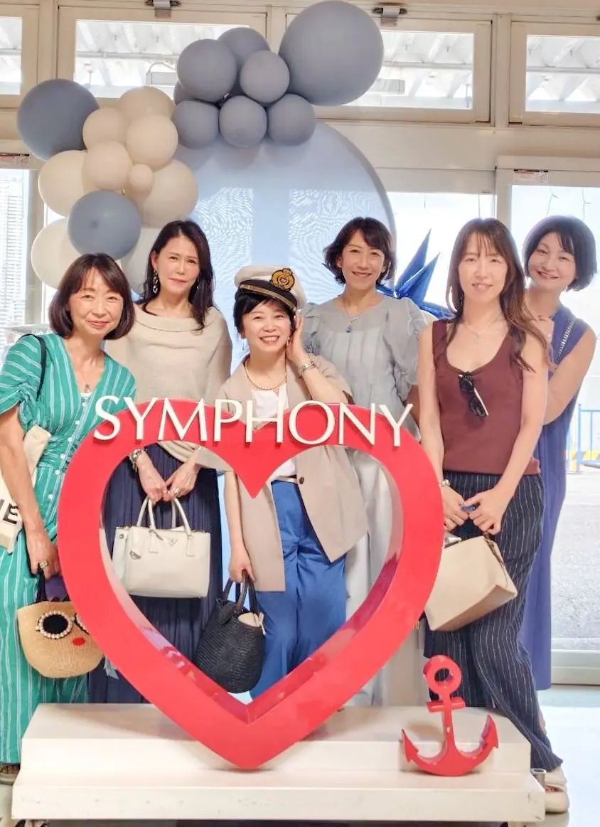 赤いハート型のオブジェを背景に、制服を着た女性を中心とした6人の女性グループが写っている。背景にはグレーの風船が飾られ、窓の向こうに建物が見える。