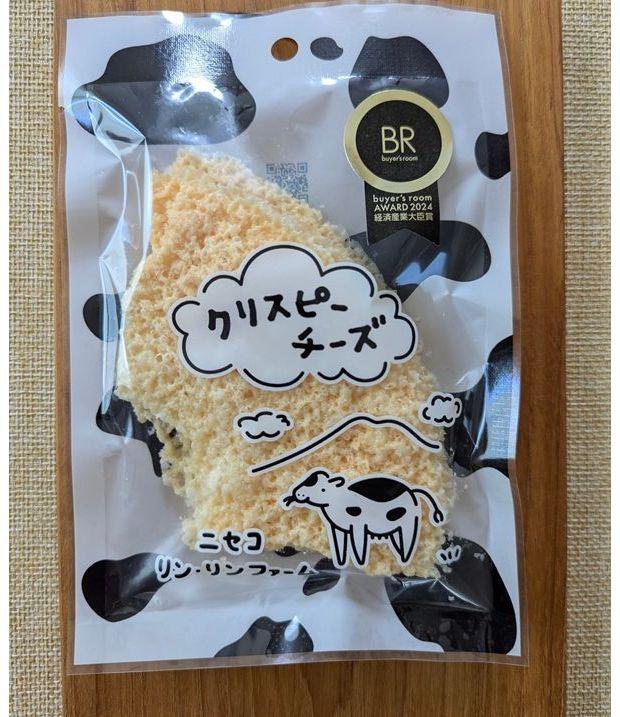牛柄のパッケージに入った、クリスピーチーズの袋。白いチーズの塊が詰まっており、「クリスピー チーズ」の文字が手書き風で描かれている。右下には牛のイラストと「ニセコ リン・リンファーム」の文字。