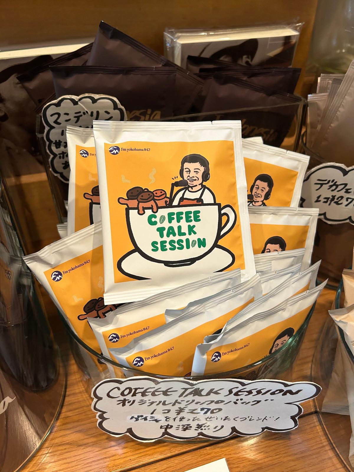 ヴィヴモン ディモンシュの「コーヒー・トーク・セッション」と書かれた黄色の個包装コーヒーパックが数個、黒い台の上に積み重ねて置かれている。パックにはコーヒーカップのイラストと、男女の顔写真が描かれている。