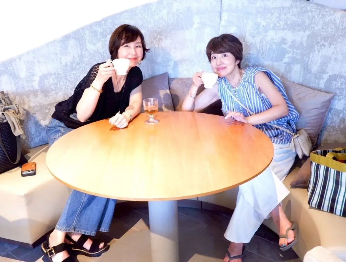 二人の女性が丸い木製テーブルに座り、コーヒーカップを手にしています。左の女性は黒のトップスとジーンズ、右の女性は青と白の縞模様のサンドレスを着ています。テーブルにはグラスに注がれたワインが置かれています。