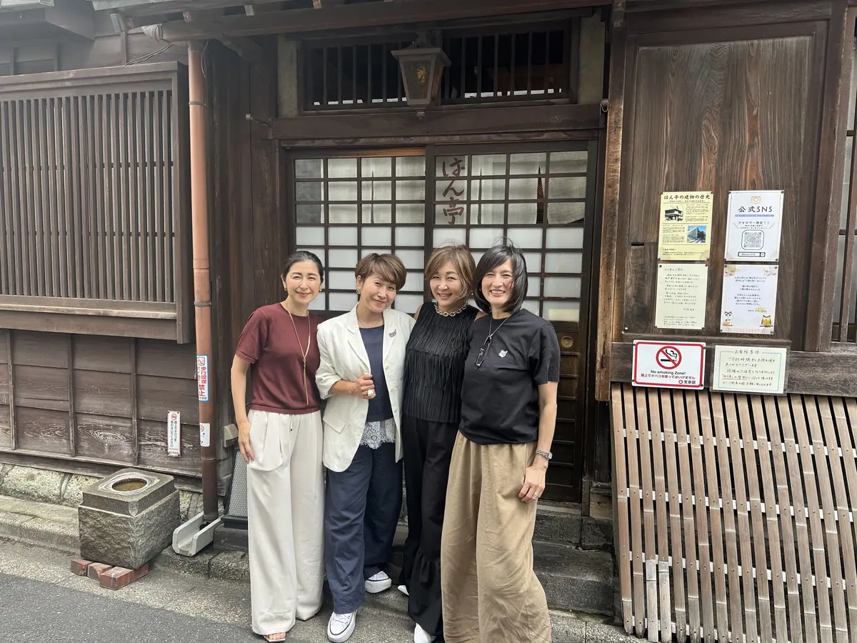 伝統的な木造建築の入り口前で、4人の女性が並んで立っている。各女性は異なる服装で、左から赤いトップス、ベージュのブレザー、黒のトップス、黒のトップスとベージュのスカートを着用。建物の壁には日本語の看板やポスターが貼り出されている。
