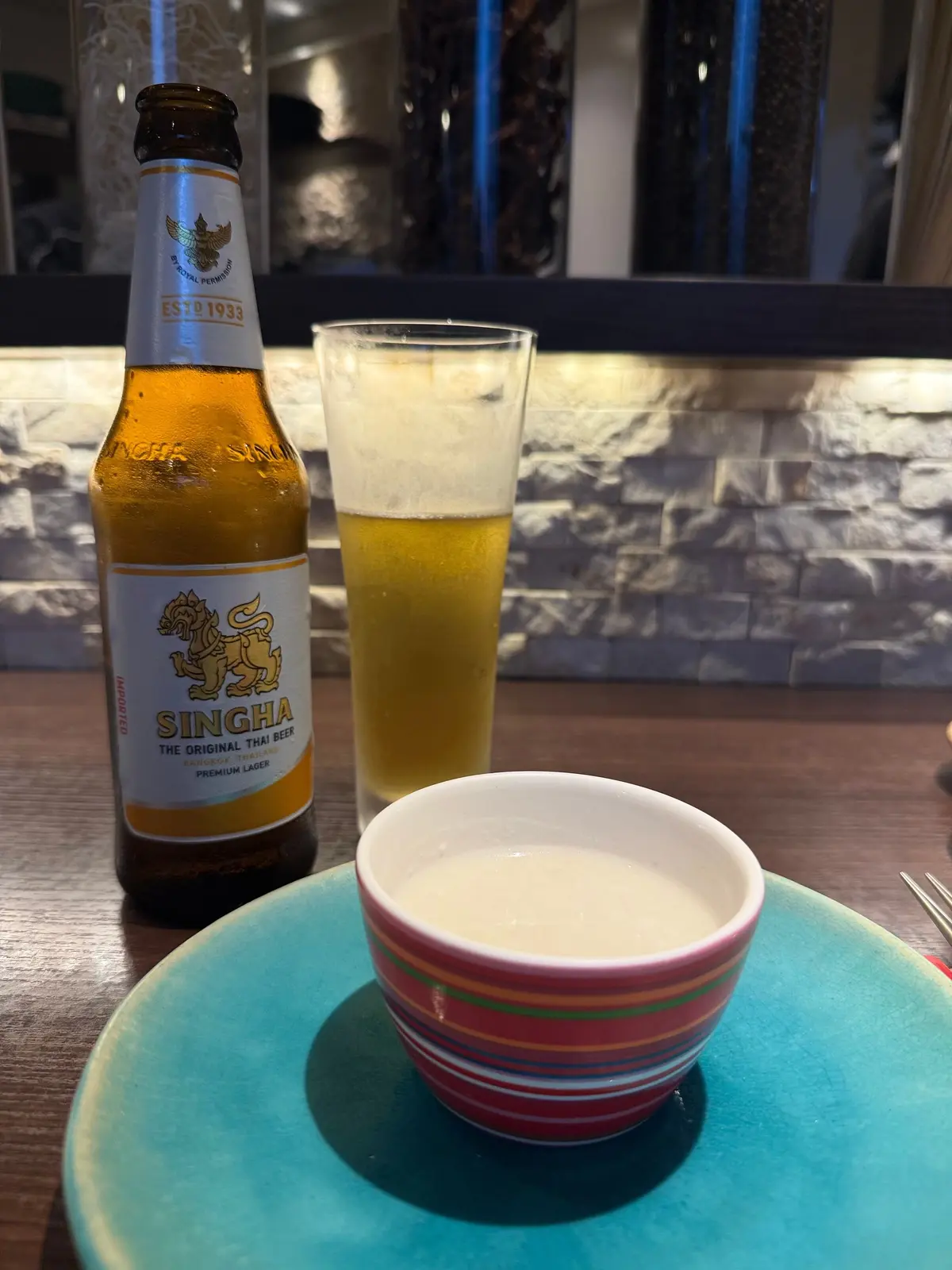 大阪・福島の「Modern Thai CIEL 大阪本店」でいただいた本格タイ料理。シンハビール石造りのテーブルに置かれた、シンハビールの一本とグラス、そしてソースを入れたカップ。ビールのボトルは黄色と赤色のラベル、グラスには泡が立っている。カップは赤、白、緑のストライプ模様。