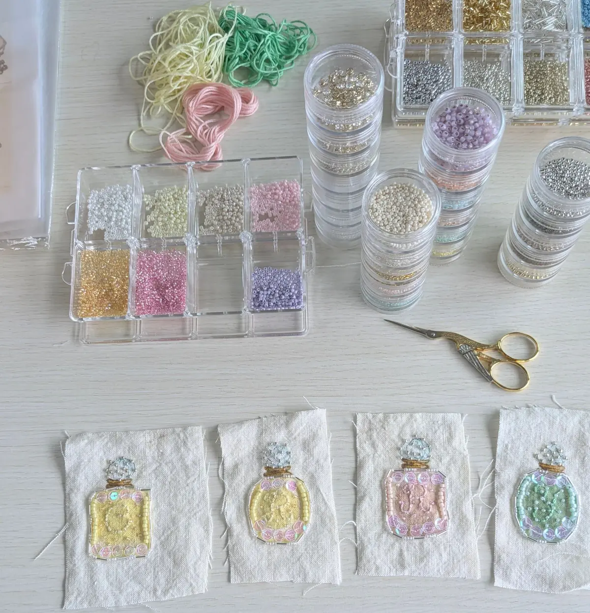白いテーブルに、ビーズや刺繍糸、ハサミが置かれた手芸のセット。様々な色と種類のビーズが容器に分かれてあり、下にはビーズ刺繍で装飾された香水の瓶のモチーフが4つ並んでいる。