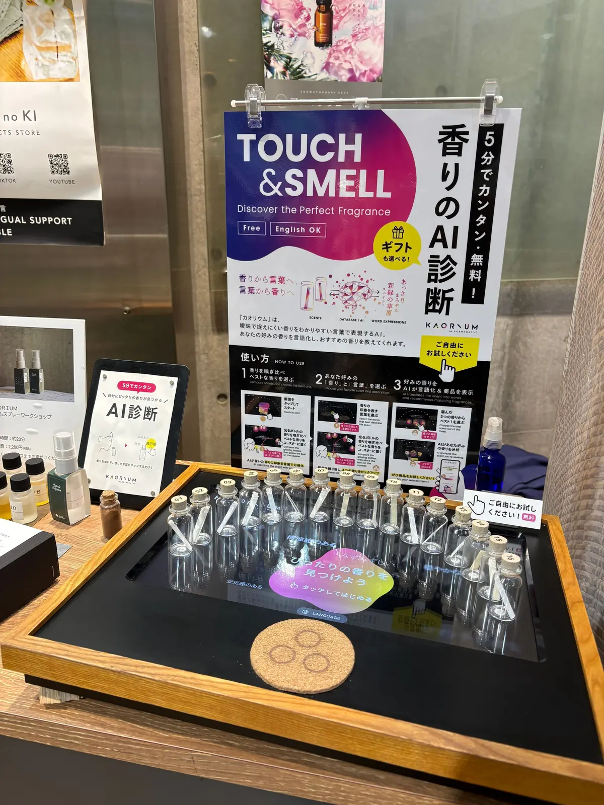 香水サンプルのボトルが並んだ木製トレイで、中央にタッチパネルと説明パネルがあり、AI香り診断の案内が書かれている