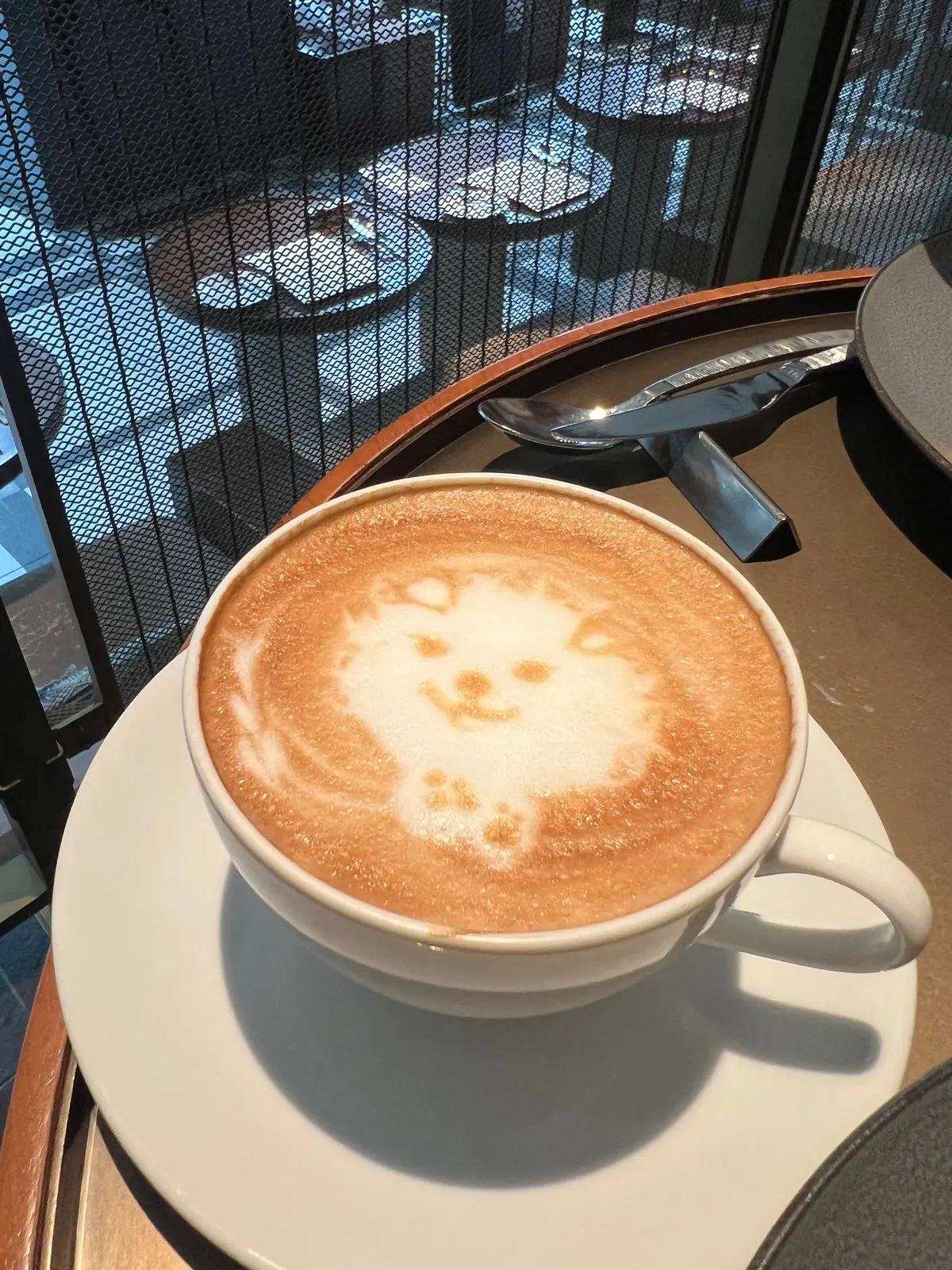 白いカップに入ったラテ。表面には、ミルクフォームで可愛らしい猫の顔の模様が描かれている。木製のトレイに置かれ、背景にはカフェのテーブルと椅子が見える。