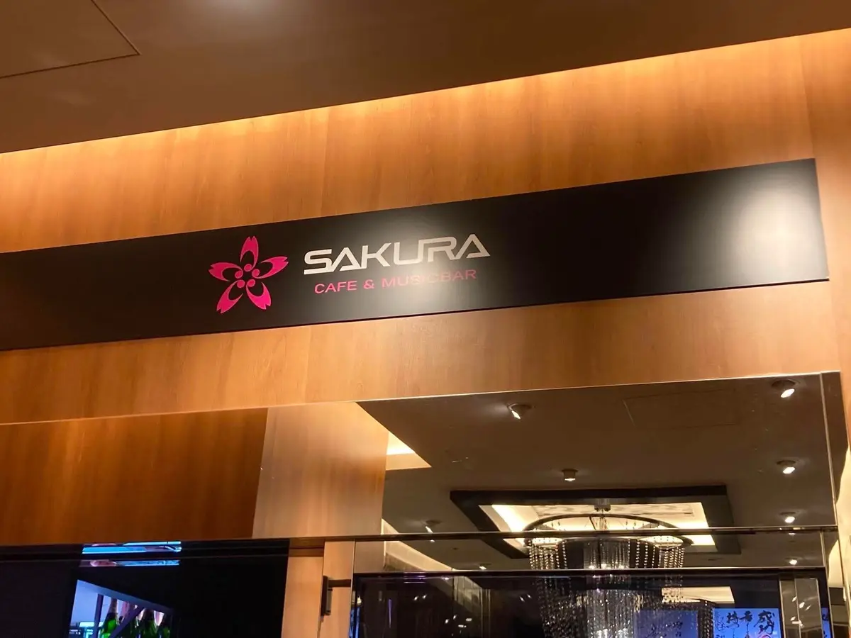木製の壁に設置された「SAKURA」の看板で、ピンクの花のロゴが特徴。ガラスの入口にはシャンデリアが吊るされ、明るい内装がちらりと見える。