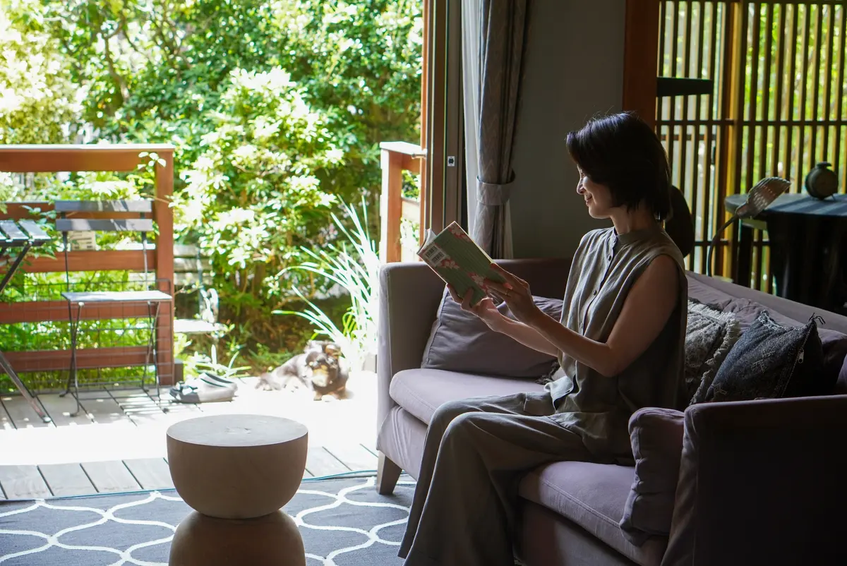 リビングルームのソファに座り本を読む女性で、窓の外には緑豊かな庭が広がる