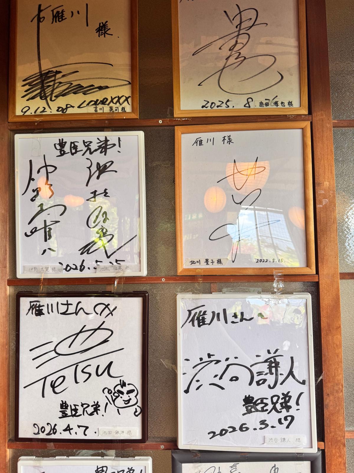 木枠に収められた白い紙に、黒いペンで手書きのメッセージが並ぶ様子。右上に「右雁川様」、左上に「書」と書かれたものなど、様々な文字やサインが書かれている。背景にはぼやけた照明が確認できる。