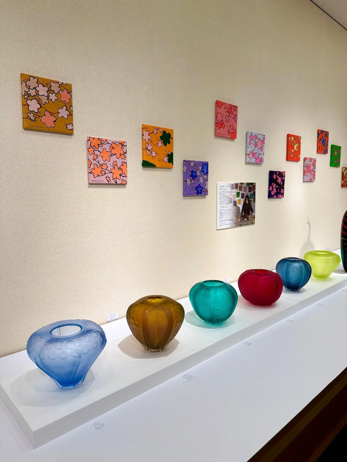 展示会場の一角。白い棚にカラフルなガラス製の花瓶が並び、背景には花柄の絵画が複数掛けられている。花瓶は青、黄、緑、赤など様々な色で、絵画はピンク、オレンジ、紫などの色調。土田康彦の個展、僕が僕であるために。あべのハルカス　ヴェネツィアンガラスアーティスト