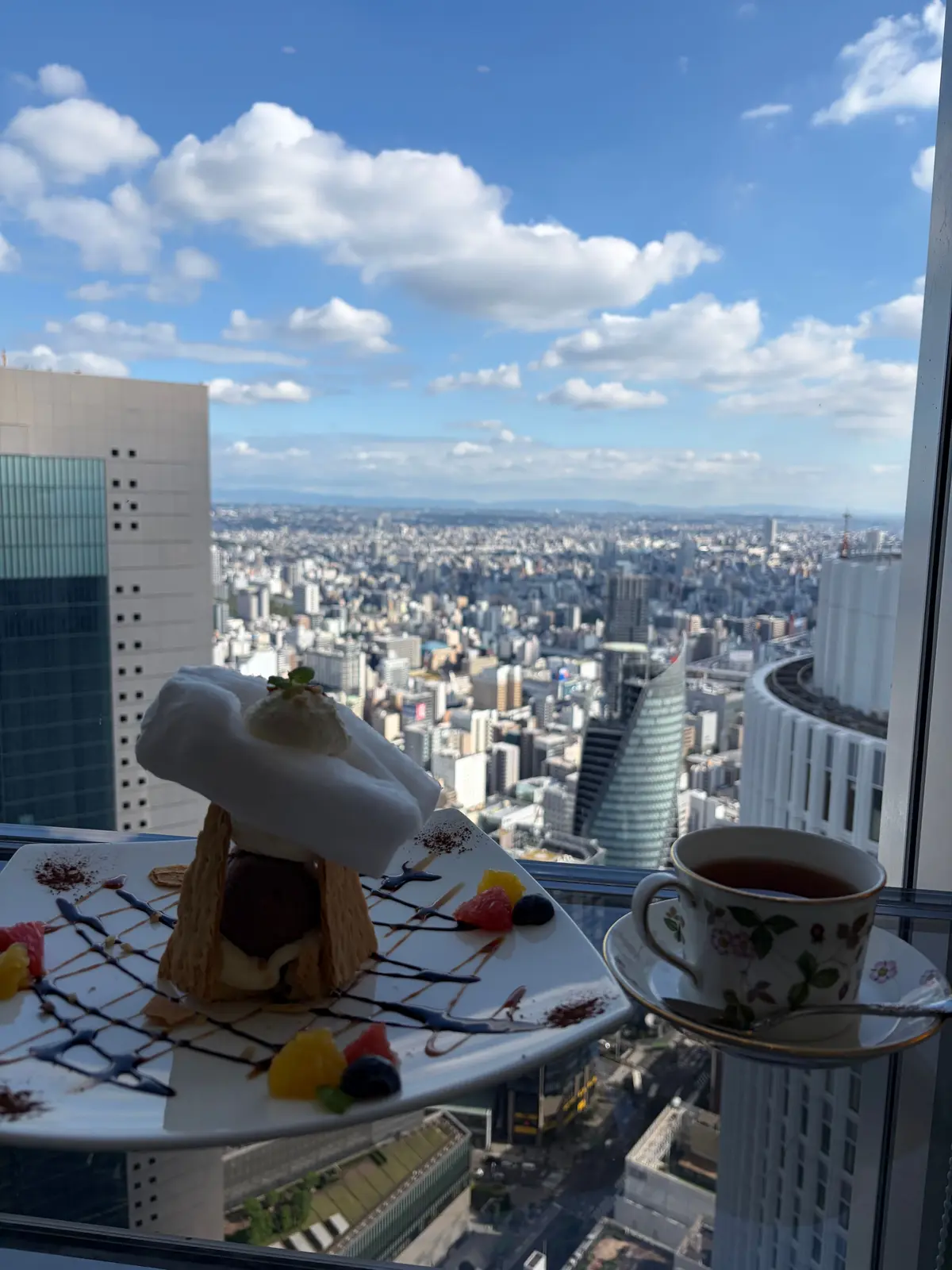 高層ビルの窓から見下ろす東京の街並み。手前に、生クリームとフルーツを添えたケーキと、花柄のカップに入ったお茶が置かれている。青空に白い雲が浮かぶ。