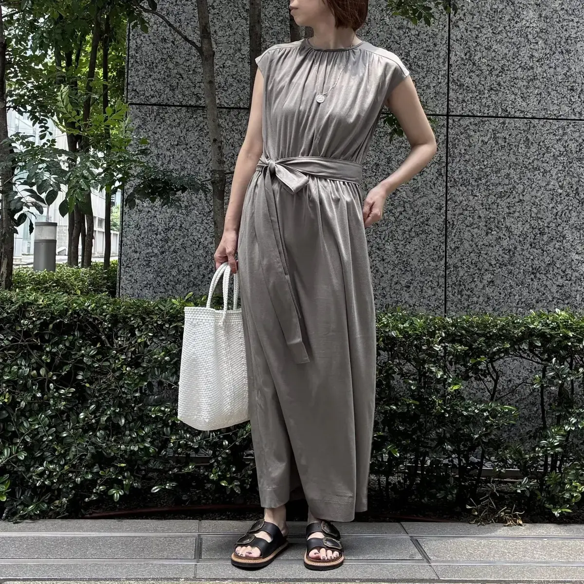 グレーのノースリーブサロペットワンピースを着た女性。腰にベルトがあり、足元は黒いサンダル。白のバッグを持ち、背景には緑の植栽とコンクリートの壁。