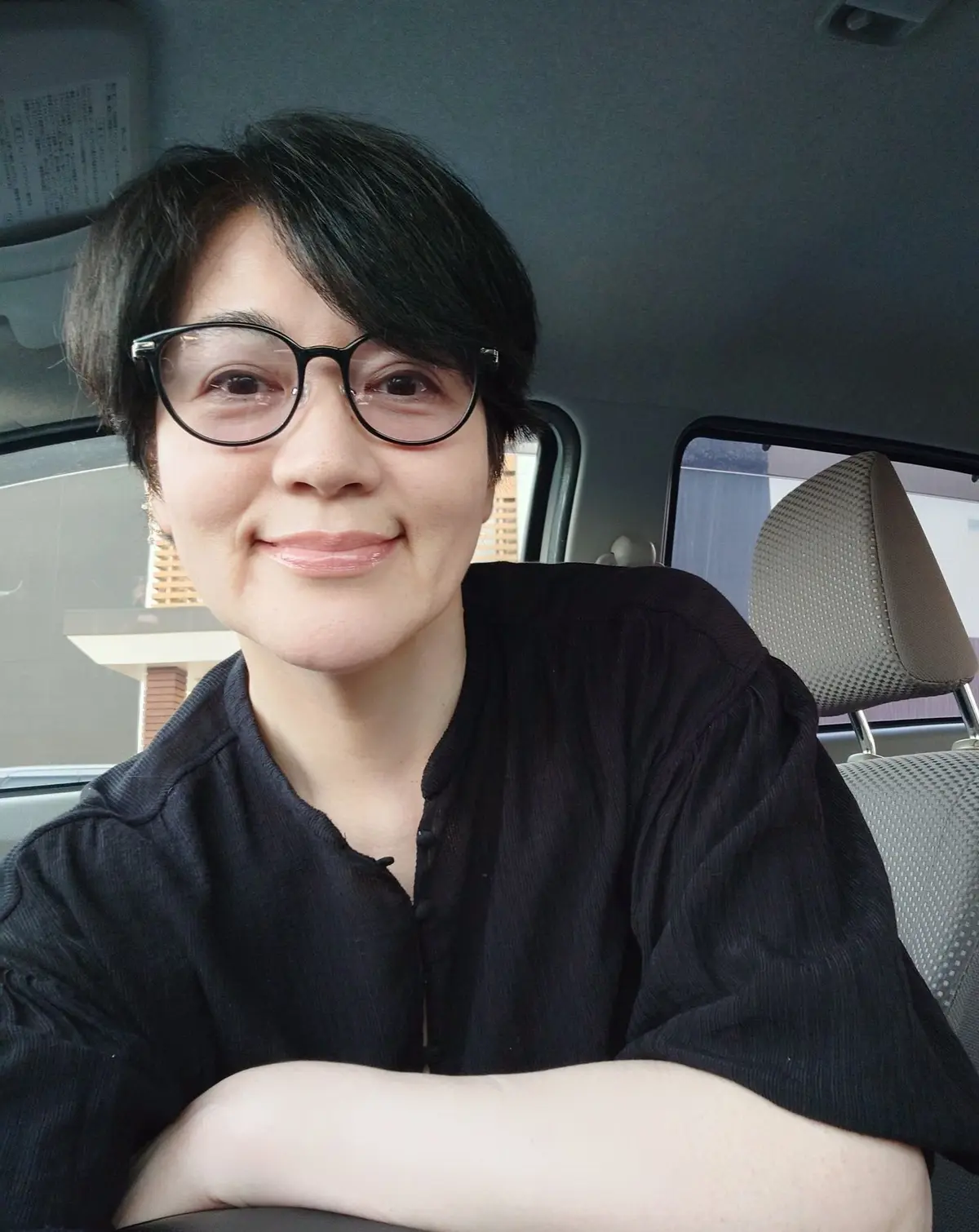 黒いフレームの眼鏡をかけた女性が、車の中で微笑んでいる。短くカットされた黒髪で、黒いシャツを着用。背景は車の内装。