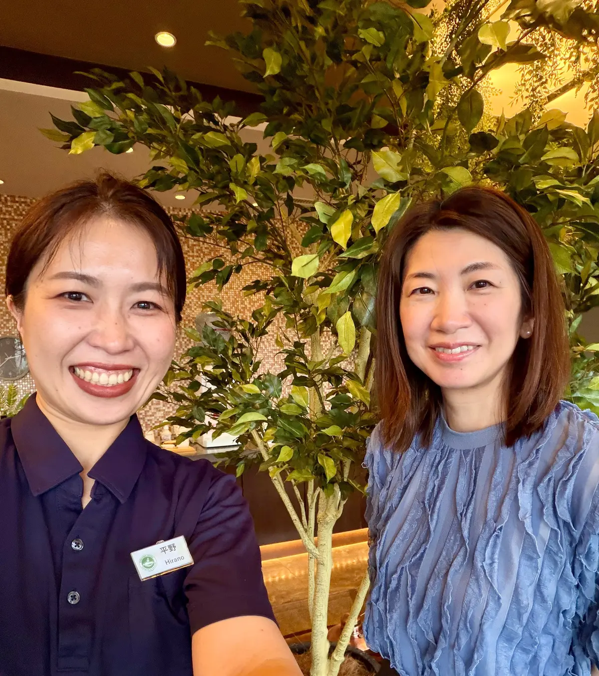 笑顔の女性2人。左は紺の制服姿のエステティシャン平野さん。右は青いフリル付きトップス。背景には緑の木と装飾照明が見える。施術後の右の女性は血行が良くなり頬が赤らんでいる。