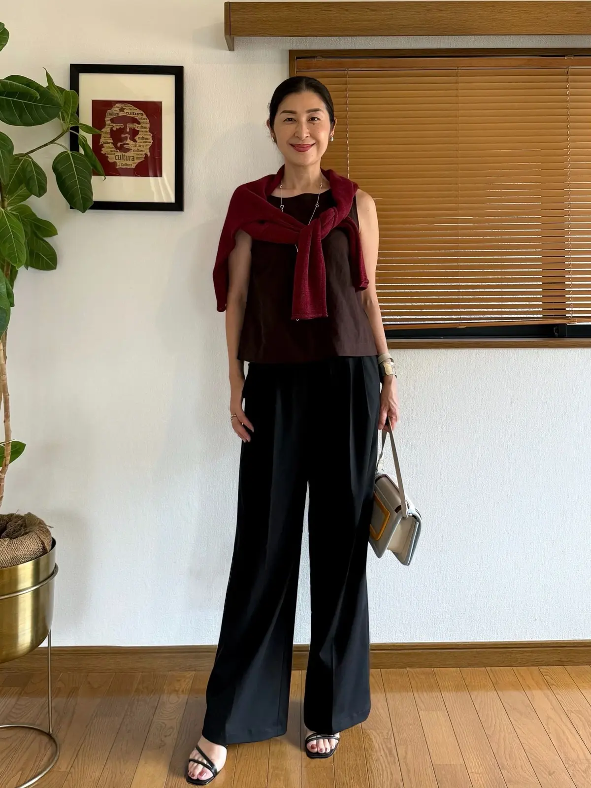 UNIQLOスマートワイドストレートパンツ、黒いワイドパンツに、ワインレッドのタンクトップとショールを羽織った女性。右手にベージュのバッグを持ち、黒いサンダルを履いている。背景には観葉植物と窓がある。