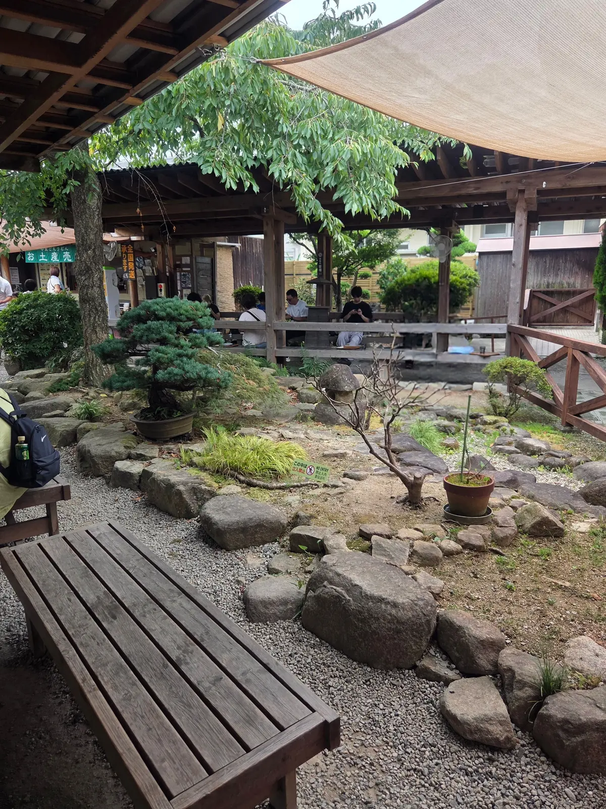 木陰の休憩所、石組みの庭園、ベンチ、テーブル席、緑の植物、屋根付きの建物。