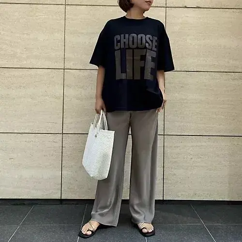 黒い「CHOOSE LIFE」ロゴTシャツ、ベージュのワイドパンツ、黒いサンダルを着用した女性。白いトートバッグを持ち、ベージュのタイル壁の前で立っている。