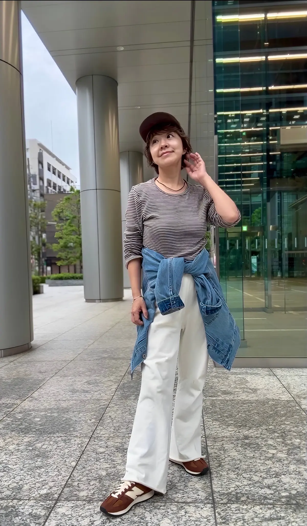 帽子を被った女性が、白いパンツとボーダーのトップス、デニムのジャケットを着用し、New Balanceのスニーカーを履いて立っている全身写真。背景には現代的な建築物。