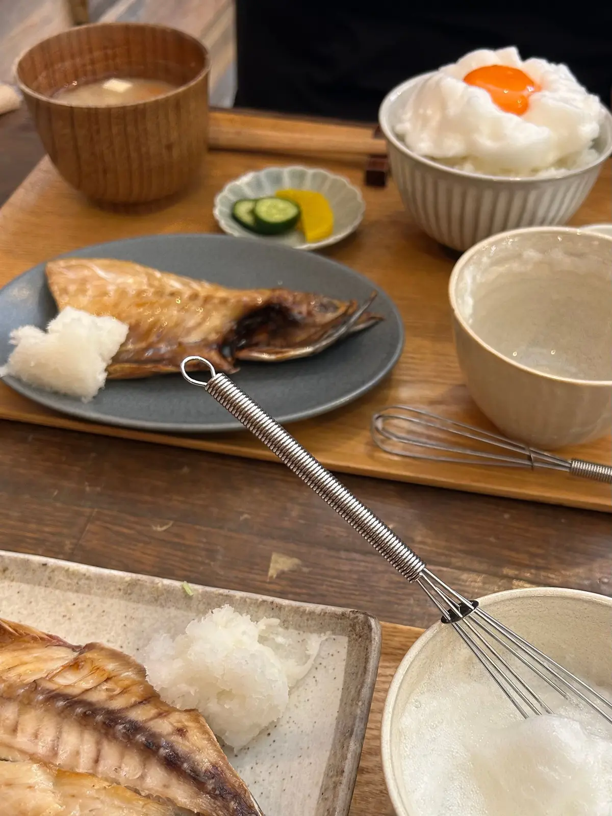 木製のトレイに盛り付けられた和食。焼き魚、ご飯、味噌汁、卵焼き、漬物が並び、泡立て器とトングが置かれている。