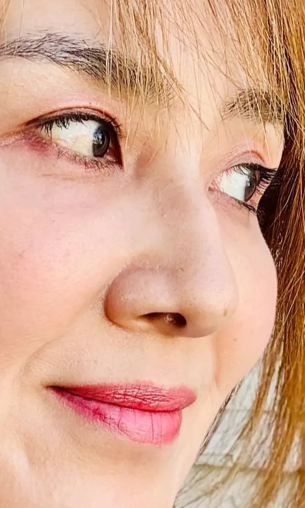 明るい茶色の髪の女性が、右目を見開いて微笑んでいるポートレート。白い花柄のイヤリングを着用し、赤いリップを付けている。青色の服を身に着けている。