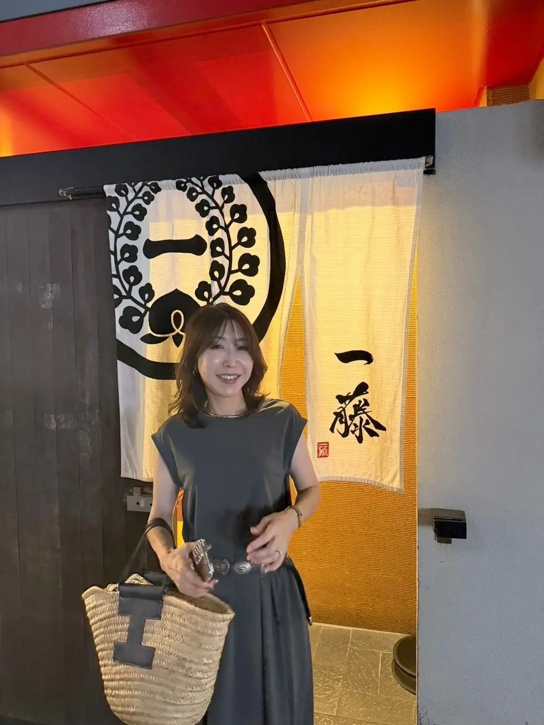 笑顔の女性が、黒いワンピースを着て立っている。背景には、黒と白の植物のイラストと、日本語の文字が書かれた白い暖簾がかかっている。天井は赤とオレンジ色に照らされている。