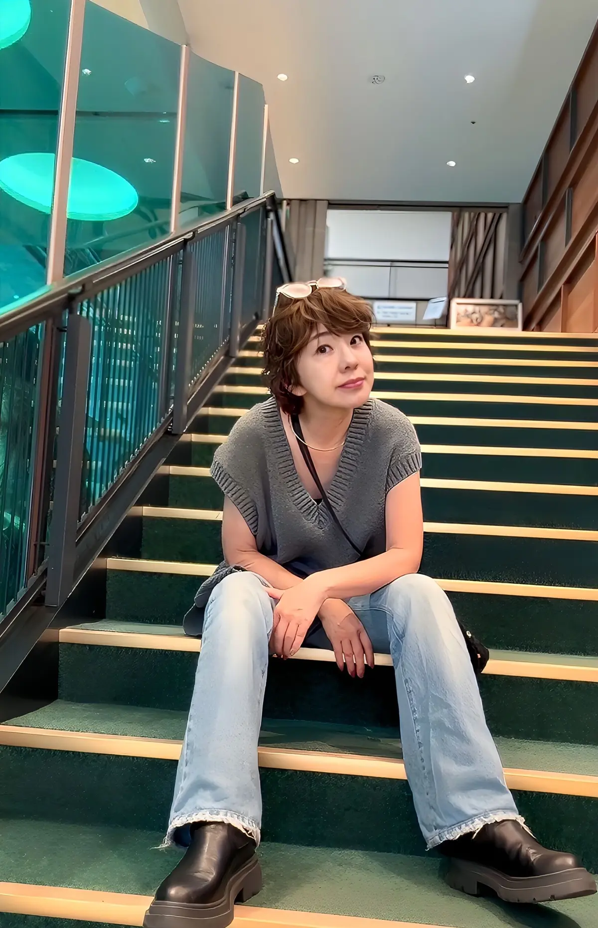 階段に座って微笑む女性。茶色のボブヘアに、グレーのベスト、ブルーのデニム、黒い厚底ブーツを着用。背景にはガラスの窓と手すりが見える。