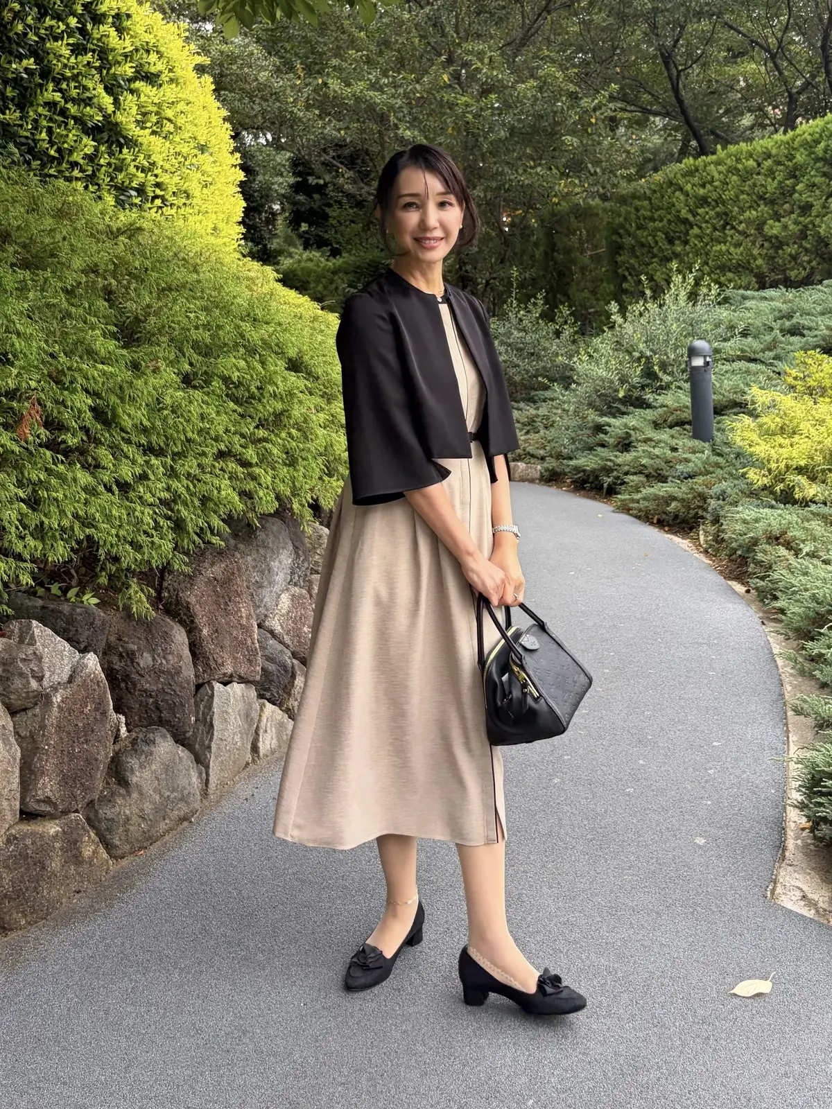 ベージュのワンピースに黒のボレロを羽織った女性が、公園の遊歩道で微笑んでいる。黒いパンプスとハンドバッグを身につけ、上品な装い。背景には緑豊かな植栽が見える。