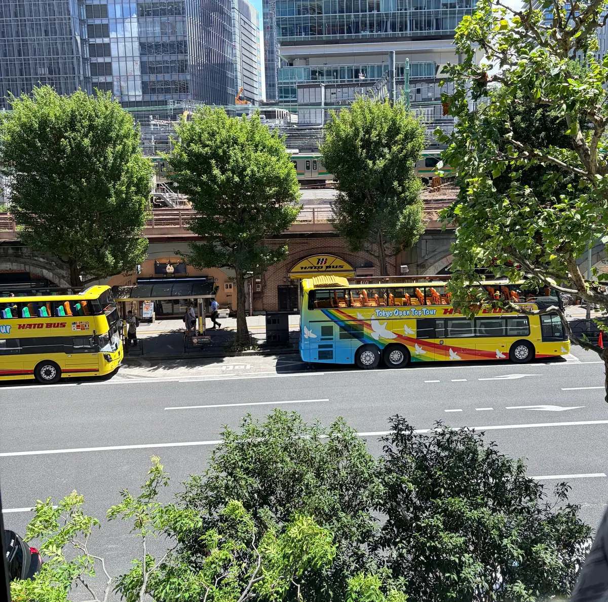 東京の街並み。道路には黄色と青色の二階建て観光バスが停車し、乗客を乗せている。背景には高層ビル群と緑豊かな木々が見える。