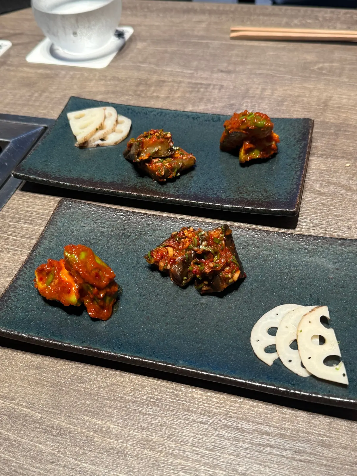 木製のテーブルに置かれた2枚の青い長方形の皿。各皿には、白く薄切りされた根菜と、赤や緑色のキムチが数点ずつ盛り付けられている。背景にはグラスと箸が見える。