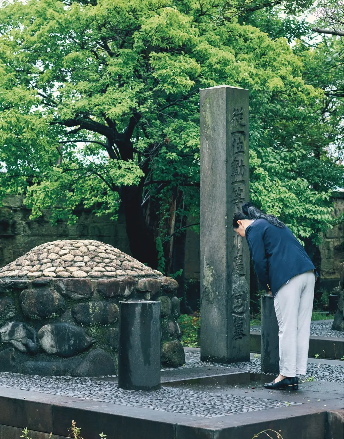 最後の将軍家が選んだ、新しい承継のかたち　徳川慶喜家の家じまい、墓じまい【前編】　緑豊かな木々の背景に、石碑と石組みの記念碑があり、若い女性が頭を下げて敬意を表している。
