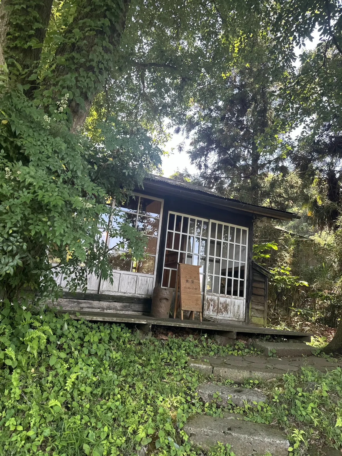 木造の小屋で、窓とドアが白い格子模様。木製の看板が入り口にあり、木製のベンチと花瓶が置かれている。木々が生い茂る自然な環境に囲まれている。