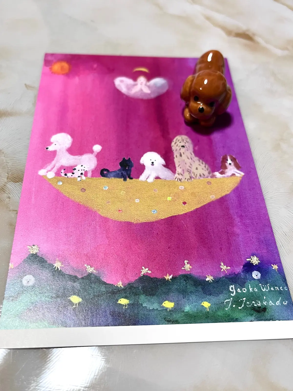 ピンク色の背景に、鳥の巣に様々な犬の置物が並んでいるイラスト。手前に黄色い鳥が数羽、下には緑の山と黄色い花が描かれている。右下に署名がある。