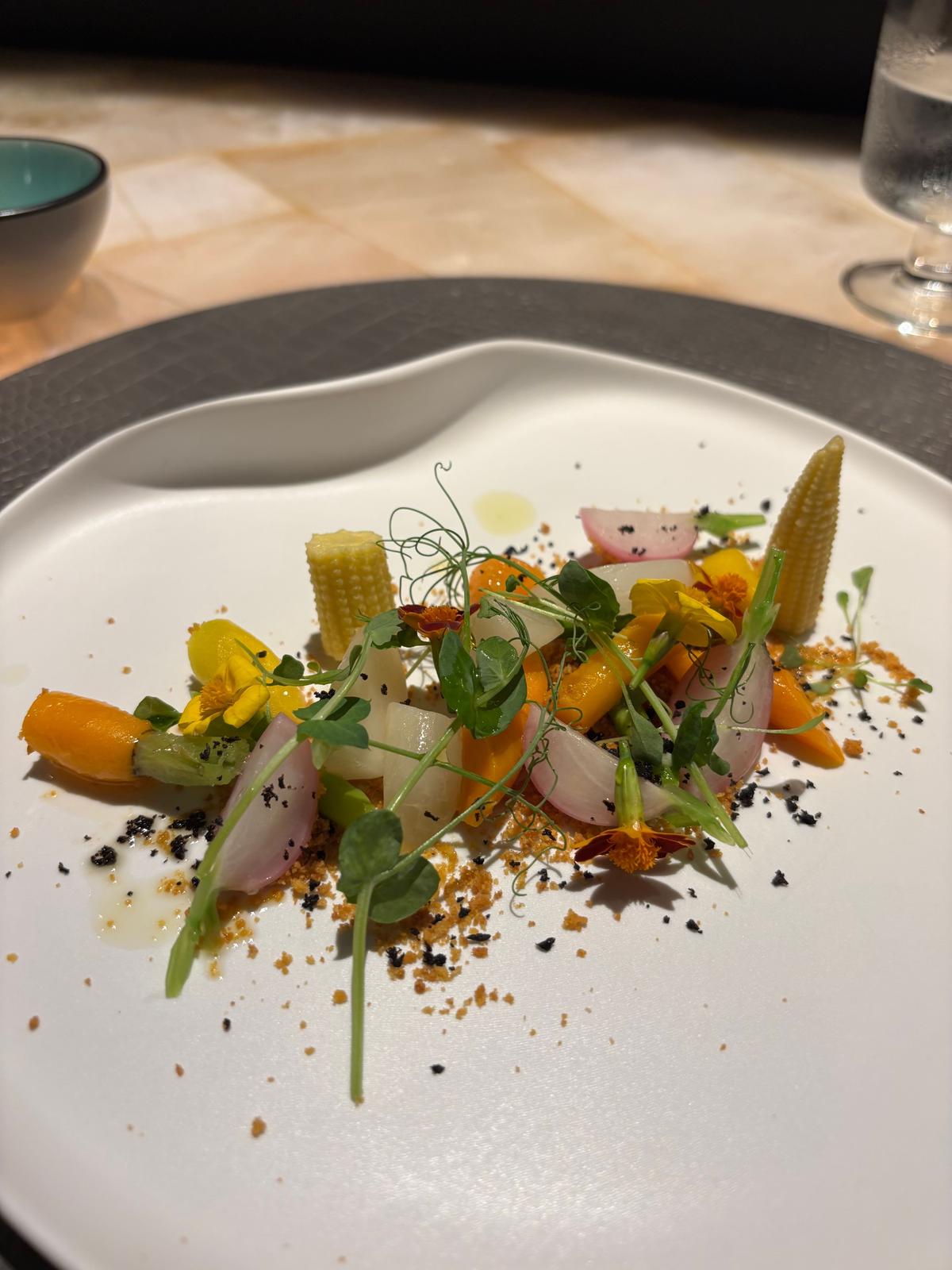 白いプレートに、黄色い花、緑のハーブ、トウモロコシの角、薄いオレンジ色の野菜、ラディッシュが美しく配置された料理。