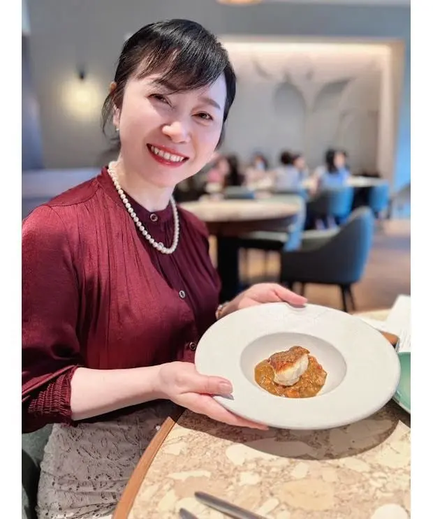 レストランで料理を前に笑顔の女性。ワインレッドのブラウスとパールネックレスを身に着け、白い皿に盛り付けられた料理を手にしている。背景には他の客席が見える。