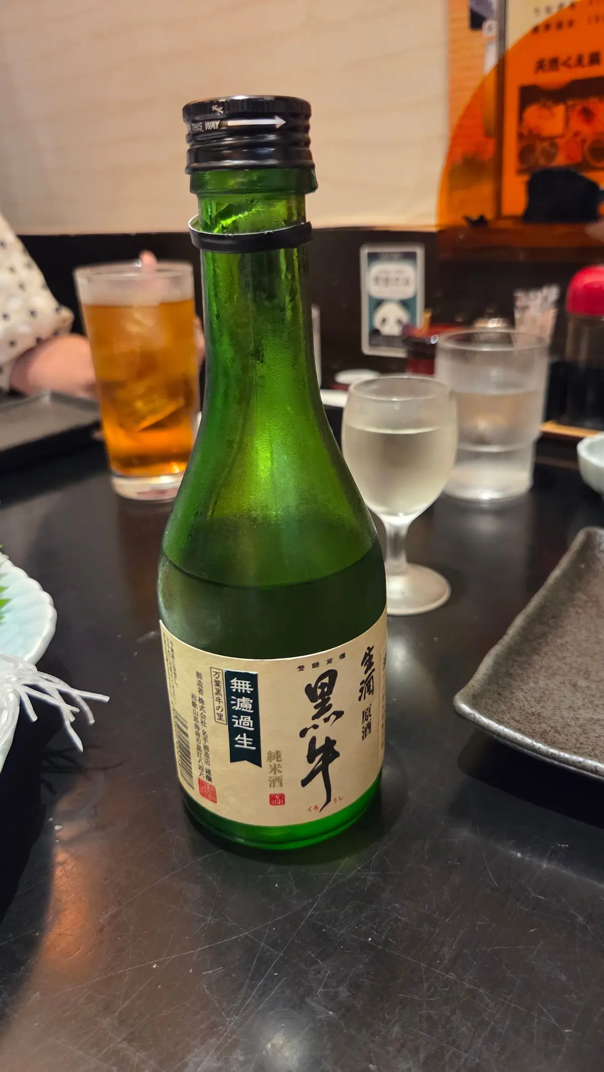 緑色の瓶に入った日本酒「男梅生」がテーブルに置かれている。近くにグラスが2つと、背景に人の手やメニューが見える。