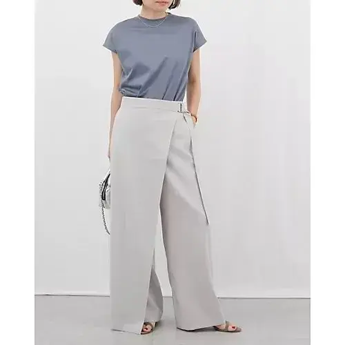 グレーのトップスとワイドパンツを着用した女性。トップスは光沢があり、パンツはフラップ付きのデザイン。背景は無地。