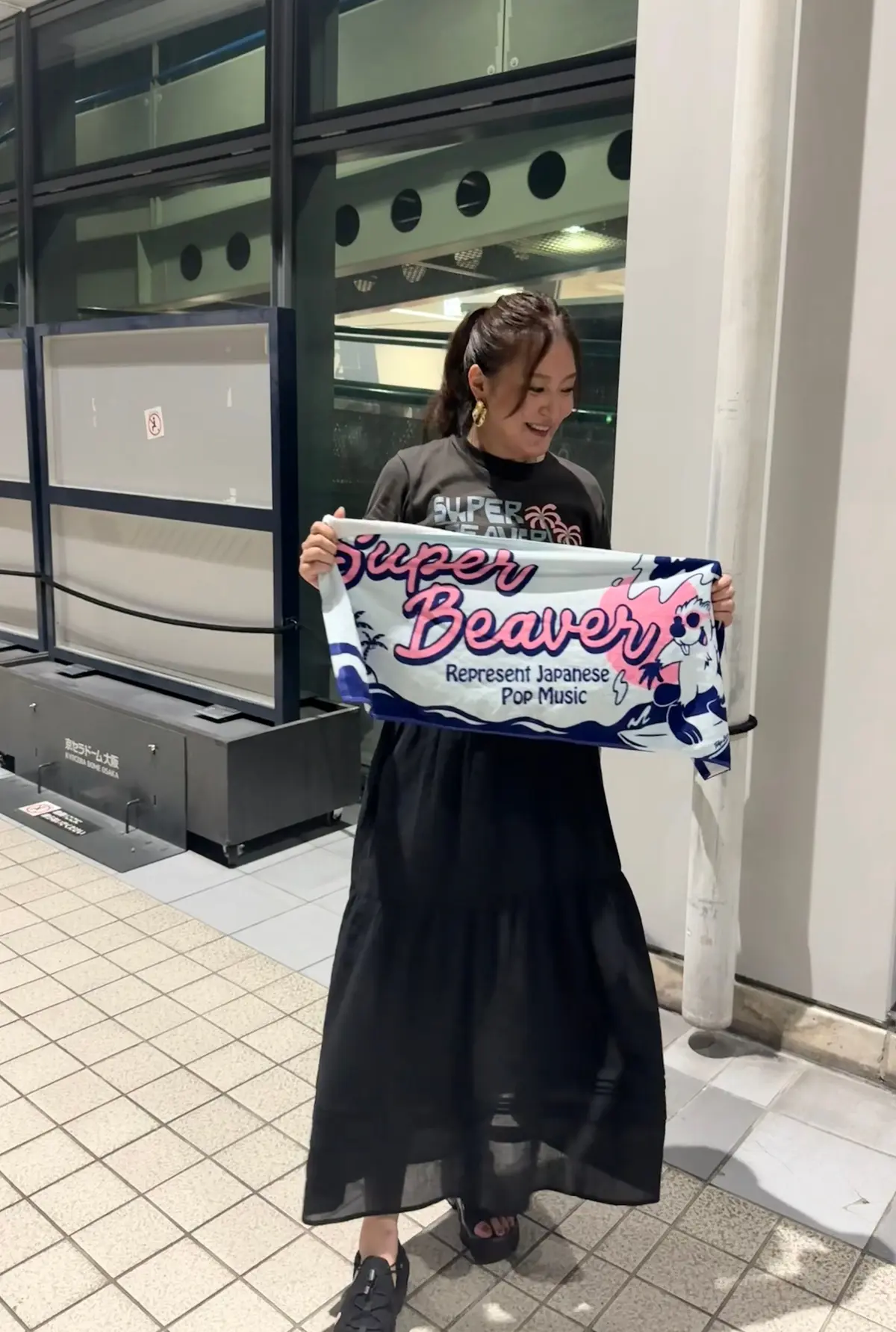 笑顔の女性が、スーパービーバー（Super Beaver）のフラッグを持ち、建物の前でポーズをとっている。茶色のロングスカートと緑色のパーカーを着用し、黒い靴を履いている。背景にはガラス張りの建物が見える。