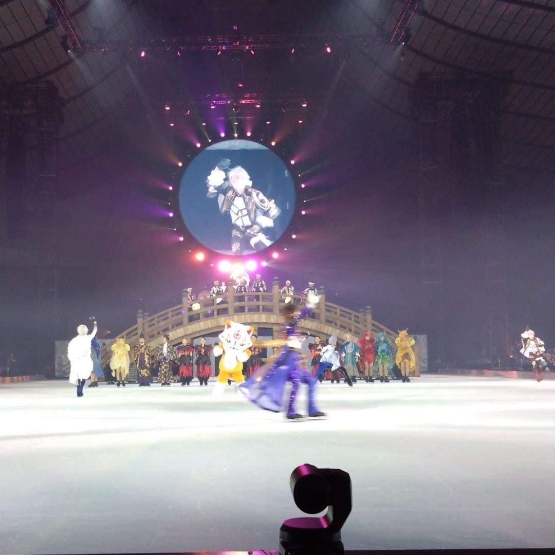 アイススケートリンクに多くのスケート選手とキャラクターが登場する公演の様子。中央には橋のセットがあり、背景には円形のスクリーンに人物が映し出されている。照明が多用され、華やかな雰囲気を演出している。