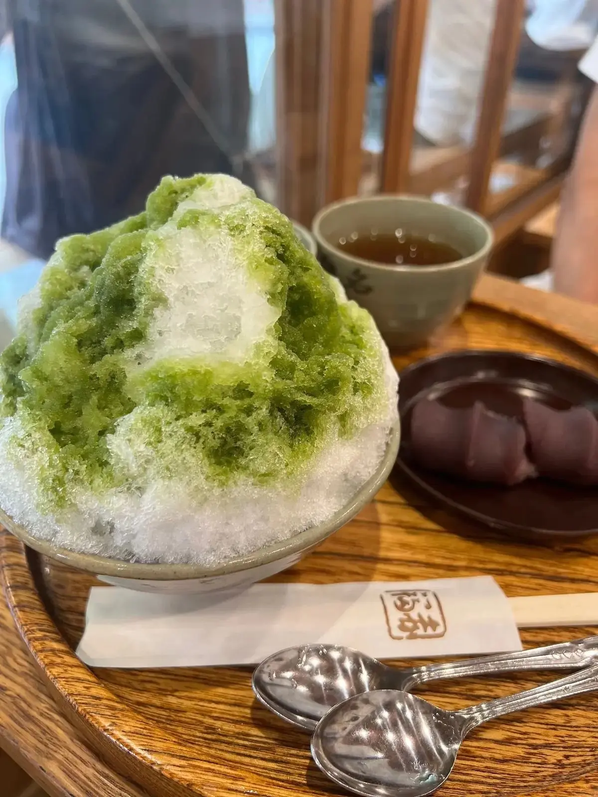 緑色のシロップをかけたかき氷が白い器に入り、木製のテーブルに置かれている。右にはお茶の入った湯呑みと、赤紫色の団子のようなものが添えられている。テーブルには紙ナプキンとスプーンが置かれている。