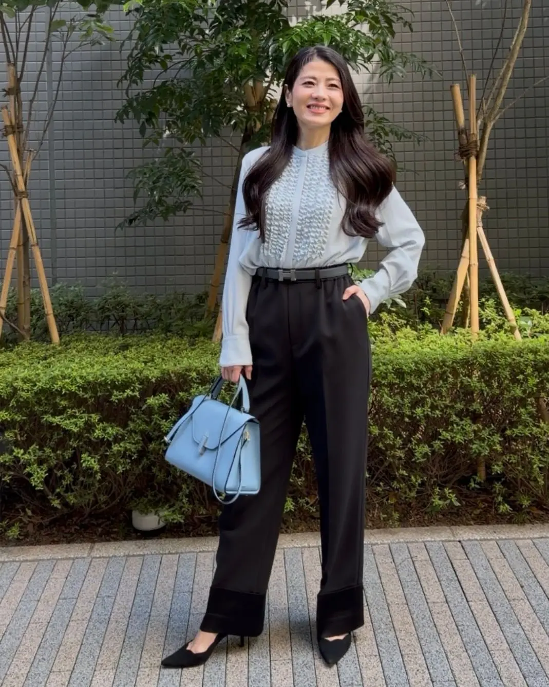 黒いハイウエストパンツと白いオーバーシャツを着用した女性が後ろを向いている。青いハンドバッグを持ち、緑の植栽エリアの前に立っている。