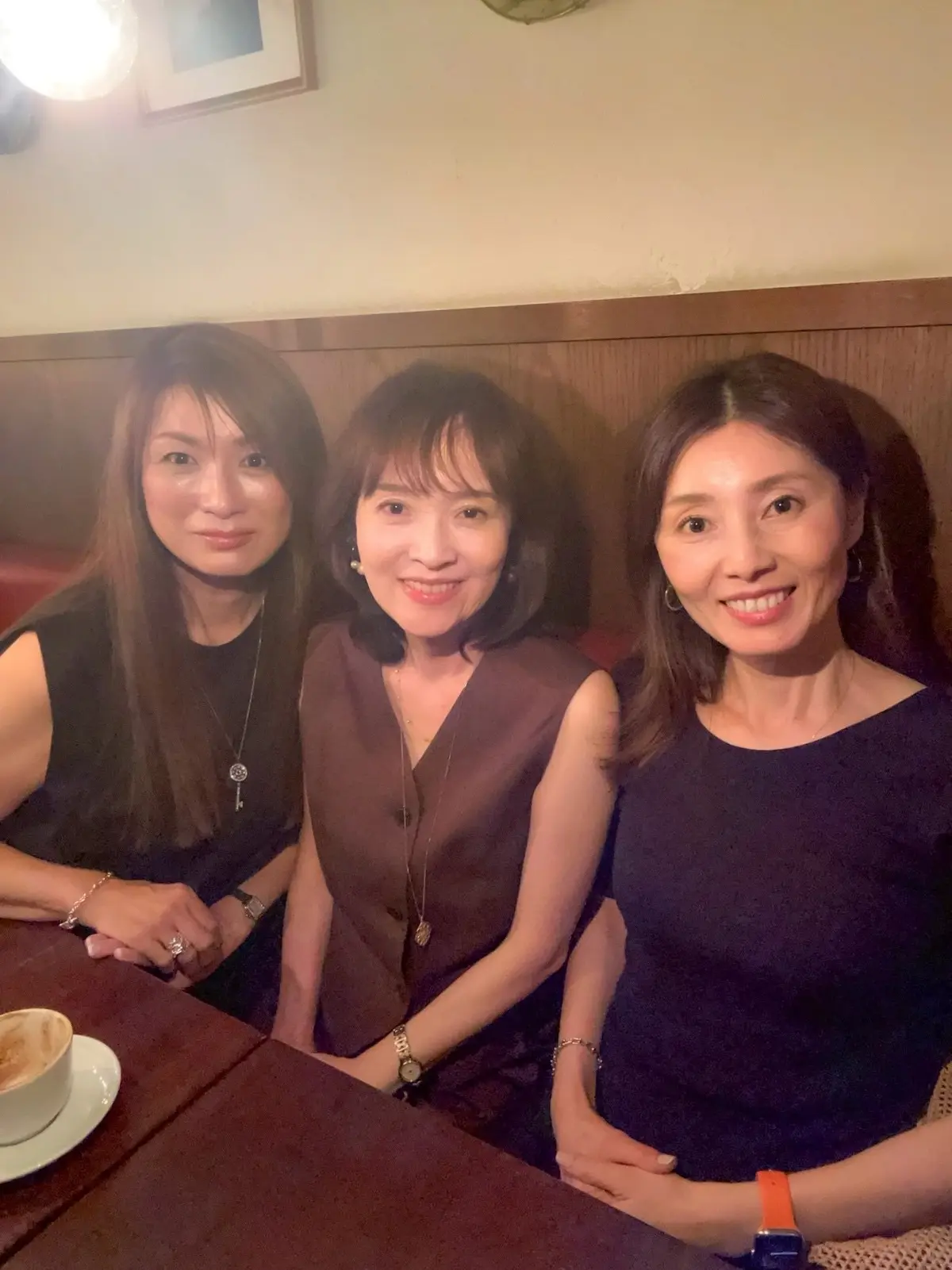 レストランで3人のアジア系の女性が笑顔で写っている。中央の女性は茶色のワンピース、左と右の女性は黒いトップスを着ている。テーブルには料理が置かれている。