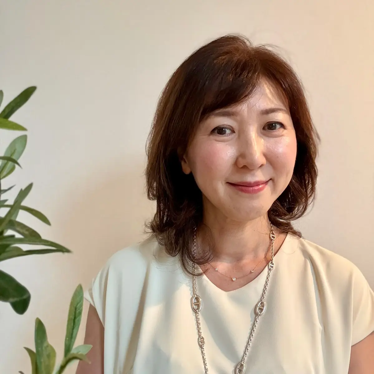 上品な笑顔の女性。茶色のボブヘアで、白いトップスを着用。ゴールドのネックレスを身につけ、背景には緑の植物が見える。