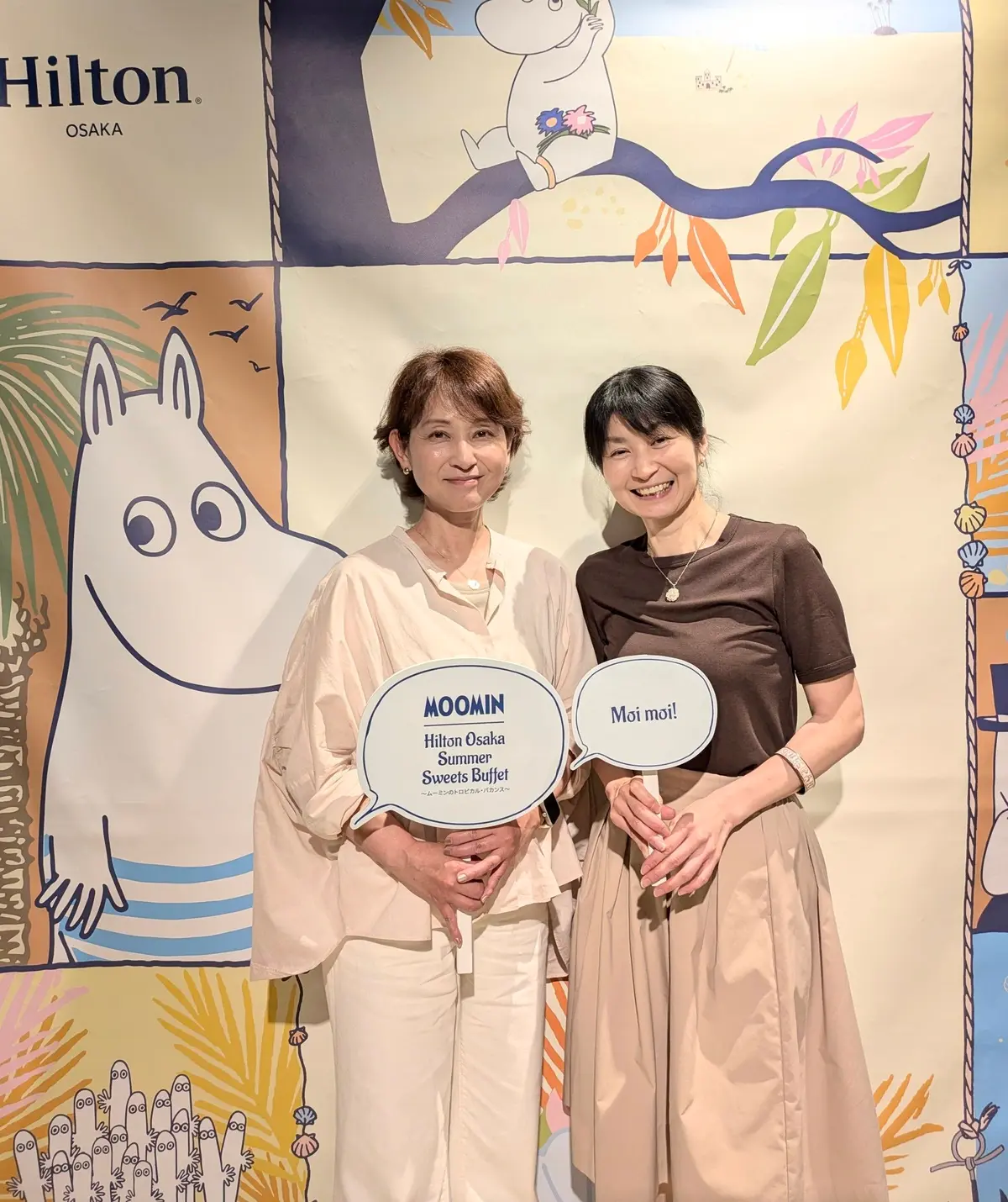 ヒルトン大阪の夏のスイーツビュッフェイベントで、ムーミンのキャラクターが描かれた背景の前で2人の女性が立ち、ムーミンのキャラクターと「Moi moi!」というテキストが表示されている