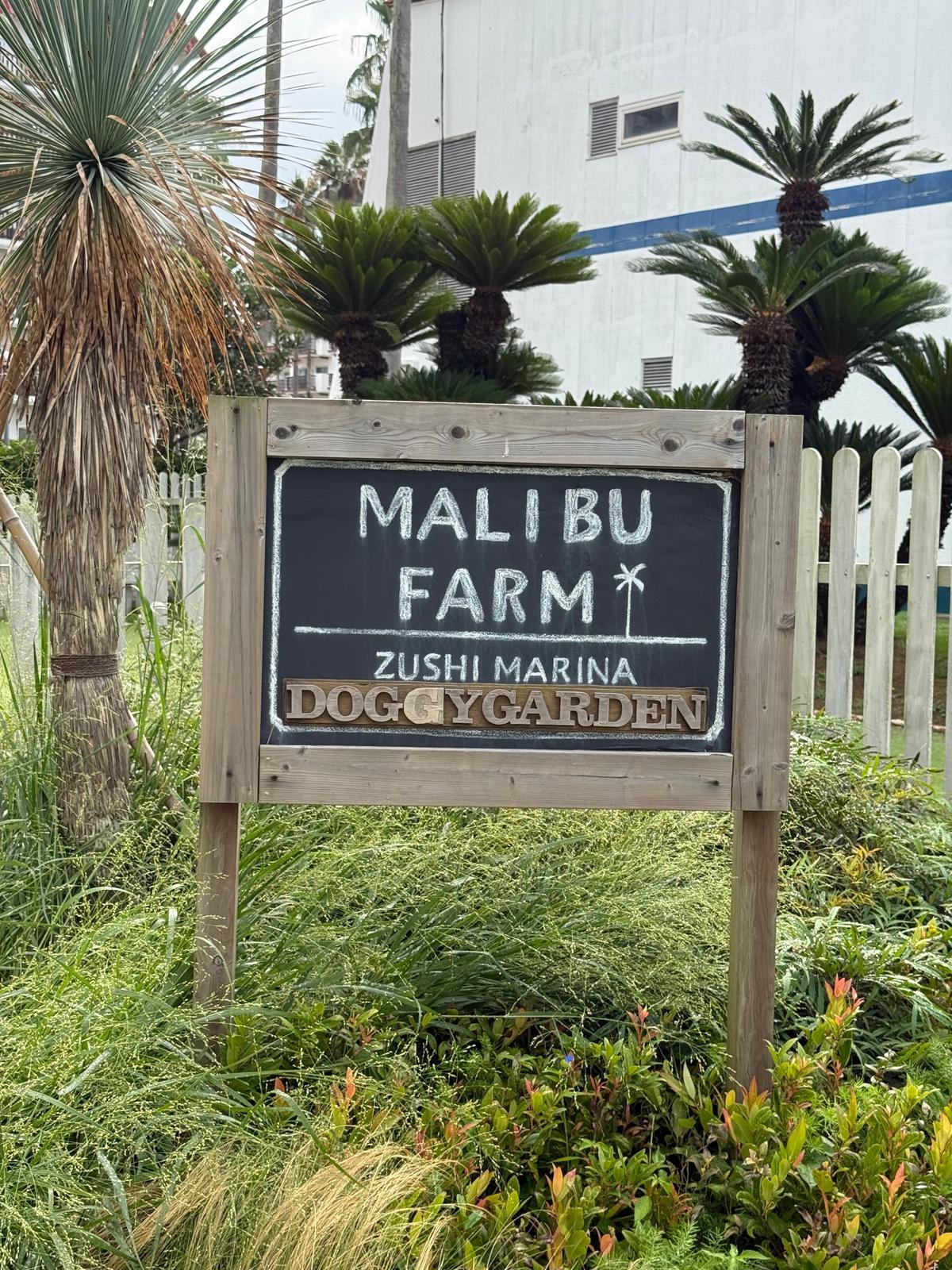 木製の看板に「MALIBU FARM ZUSHI MARINA DOGGY GARDEN」と書かれている。背景には、建物とヤシの木が見える。看板の周りは草が生い茂っている。